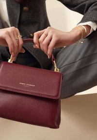 Sac à main en cuir bordeaux avec une surface texturée, quincaillerie dorée et une poignée supérieure. Dispose d'une fermeture à rabat et d'un logo de marque.