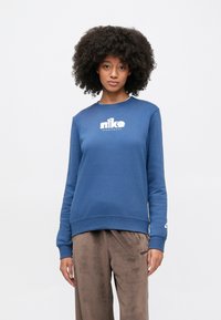 Μπλε φούτερ από βαμβακερό μείγμα με λαιμόκοψη crew, ριμπ μανσέτες και λευκό λογότυπο Nike στο στήθος. Συνδυάζεται με καφέ βελούδινο παντελόνι.