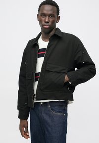 Chaqueta corta negra hecha de tela texturizada, con cremallera frontal, dos bolsillos con solapa y un ajuste casual. Lleva puesta sobre un suéter de rayas.