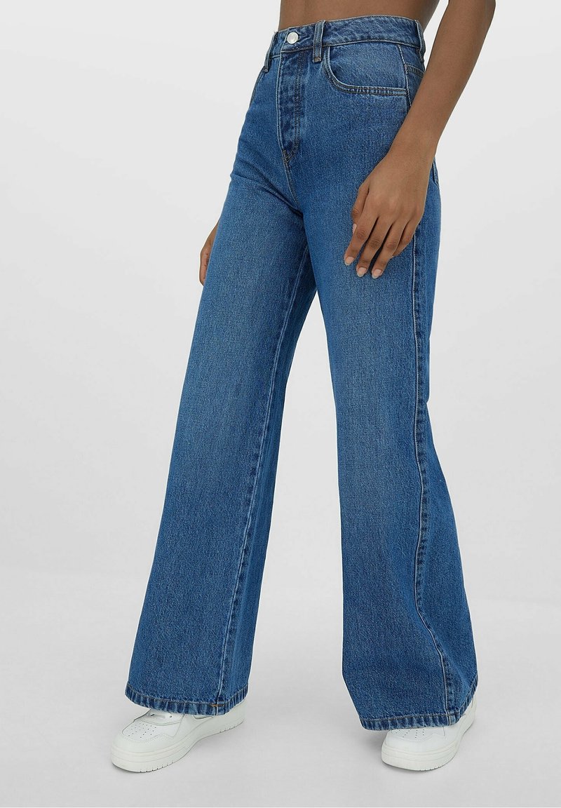 Korkeavyötäröiset, leveälahkeiset siniset farkut, joissa on hieman haalistunut pinta, etutaskut ja nappisuljenta. Yhdistetty valkoisiin tenisseihin.