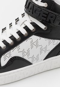 Close-up of black and white high-top sneaker with white laces, perforated star patterns, and bold brand name embossed on the sole and strap.