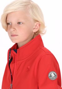 Rød softshell-jakke med høj krave, lynlås foran og logo-patch på ærmet, med en glat tekstur og en skræddersyet pasform.
