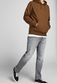 Sudadera con capucha marrón y bolsillo delantero, superpuesta a una camiseta blanca. Combinada con jeans gris claro y zapatillas negras con cordones blancos.