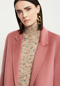 Abrigo de lana rosa claro con un diseño estructurado, combinado con un suéter de punto texturizado multicolor con toques en tonos pastel. Pendientes grandes y gruesos.