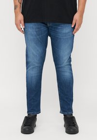 Kék farmer skinny farmernadrág, elhalv�ányult mosással, amelynek keskeny szabása és diszkrét gyűrődése van, fekete, alacsony szárú sportcipővel párosítva.