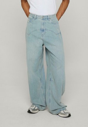 Jeans en denim bleu clair, à jambes larges, dotés d'une taille haute, de poches avant et d'une fermeture à boutons. Portés avec des baskets argentées.