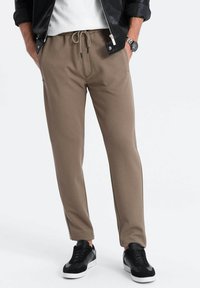 Bruine joggingbroek met een gestructureerde stof en taps toelopende pijpen, gecombineerd met zwarte sneakers, op een neutrale achtergrond.