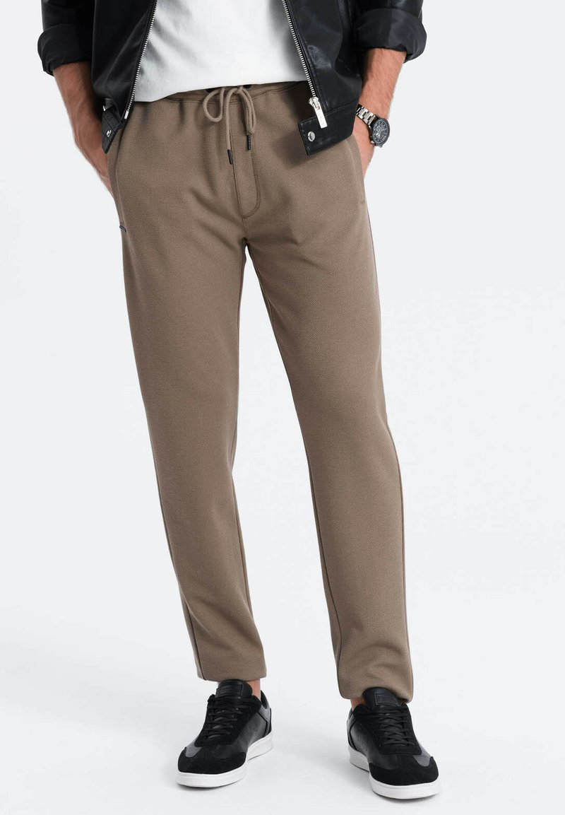 Bruine joggingbroek met een gestructureerde stof en taps toelopende pijpen, gecombineerd met zwarte sneakers, op een neutrale achtergrond.