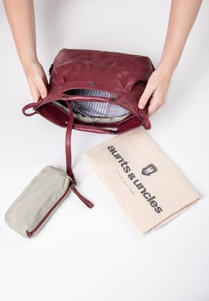 Bordeauxfarbene Ledertasche mit gefüttertem Innenraum, das ein gestreiftes Muster zeigt. Enthält eine beigefarbene Leinentasche mit der Aufschrift "aunts & uncles" und ein Reißverschlusszubehör.