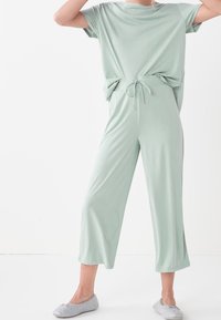 Lichtgroene jumpsuit met korte mouwen, een trekkoord in de taille, een relaxed fit en cropped pijpen. Zachte stof met een gladde textuur. Grijze slippers.