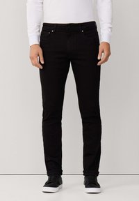 Vaqueros de mezclilla negros con un ajuste slim, diseño de cinco bolsillos y detalles metálicos. Combinados con zapatillas negras y una camisa blanca de manga larga.