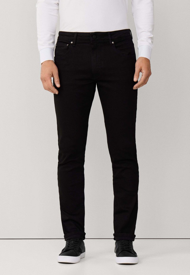 Vaqueros de mezclilla negros con un ajuste slim, diseño de cinco bolsillos y detalles metálicos. Combinados con zapatillas negras y una camisa blanca de manga larga.