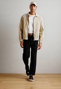 Jachetă bomber bej peste o cămașă albă, pantaloni negri și adidași negri. Modelul poartă o șapcă asortată, stând pe un podea din lemn.