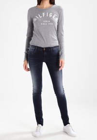 Šedé tričko s dlouhým rukávem s potiskem „HILFIGER DENIM SINCE 1985“, doplněné tmavě modrými úzkými džínami a bílými teniskami. Jednoduchý, přiléhavý střih.