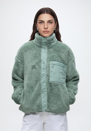 Femme aux longs cheveux foncés portant des boucles d'oreilles argentées en forme de créoles et une veste polaire vert clair avec une poche poitrine zippée et des pressions, mains dans les poches.