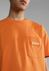 T-shirt di cotone arancione con collo a giro e taschino sul lato sinistro del petto. Presenta una toppa con logo bianco e arancione sulla tasca.