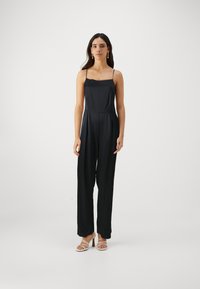 Svart ärmlös jumpsuit med rak halsringning, figurnära midja och vida byxor, tillverkad av smidigt tyg. Bärs med sandaler med remmar.