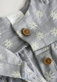 Prenda de tela gris con patrones de flores ligeros, que presenta botones de madera y una etiqueta que dice "NEXT baby." Textura suave y diseño casual.