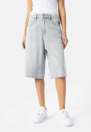 Person iført lysegrå knælange denimshorts, hvide sneakers og en hvid top, stående på en ensartet lys baggrund.
