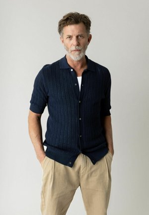 Mand med gråt skæg iført navyblå kortærmet strikket cardigan og beige bukser, stående med hænderne i lommerne mod en ensfarvet baggrund.