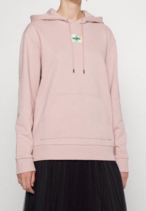 Personne portant un sweat à capuche rose pâle avec poche avant et logo « HUGO », associé à une jupe plissée noire sur un fond uni.