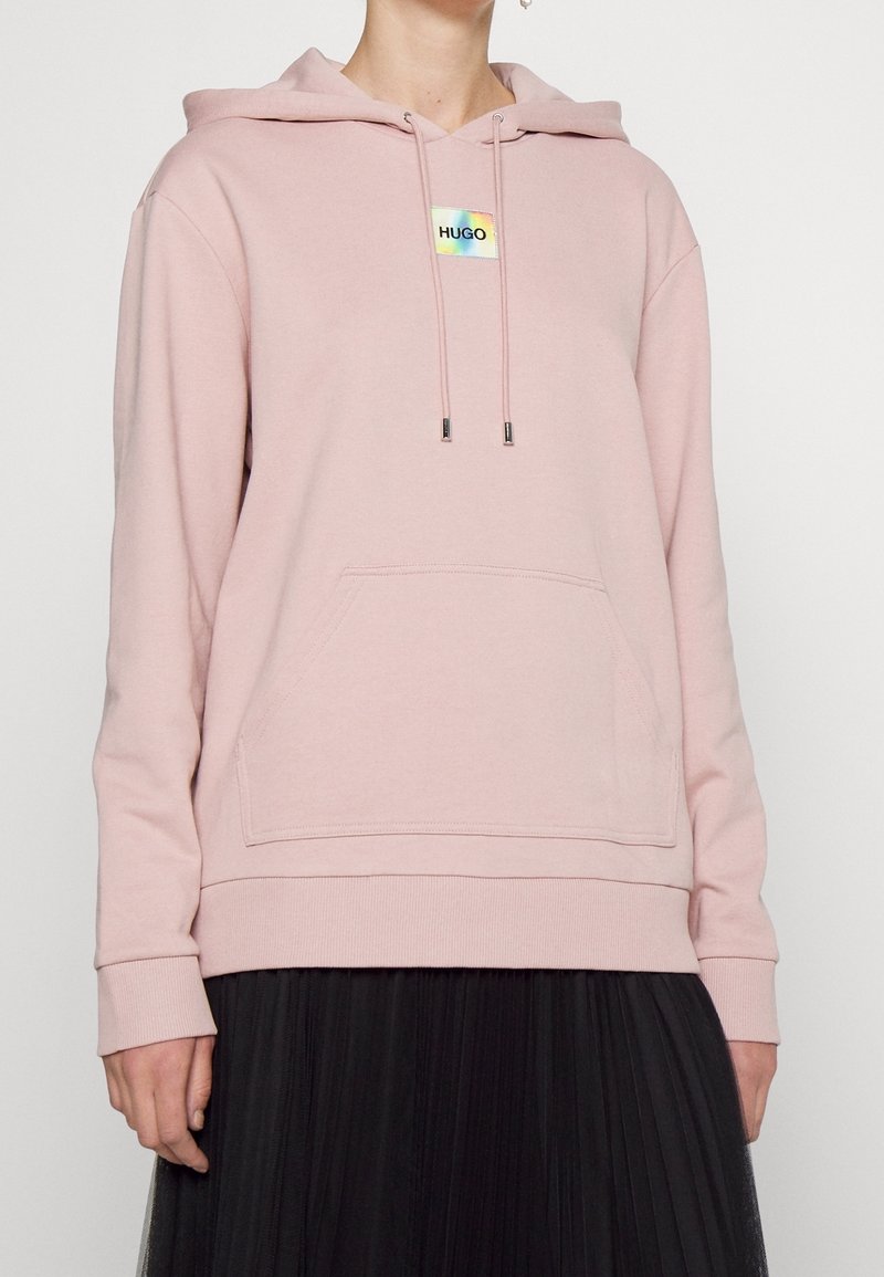 Personne portant un sweat à capuche rose pâle avec poche avant et logo « HUGO », associé à une jupe plissée noire sur un fond uni.