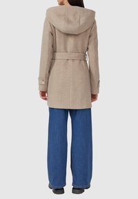 Manteau en mélange de laine beige avec une capuche, ceinture à la taille et poignets boutonnés, porté sur un jean en denim bleu à jambes larges. Texture lisse, design ajusté.