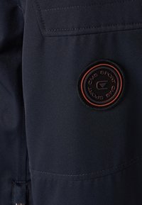 Mörkblått tyg med en slät textur, med en rund svart och röd logotypapplikation. Inkluderar synlig sömnad och en sidofickdesign.