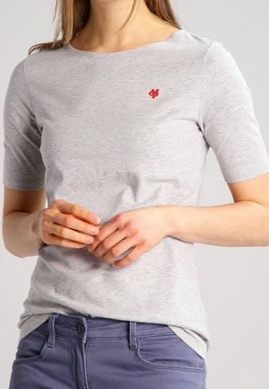 Person trägt ein hellgraues kurzärmeliges Hemd mit einem kleinen roten Logo auf der Brust und blaue Hosen, die Hände sanft vor der Taille verschränkt.