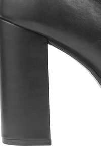 Zapato de tacón alto de cuero negro con un tacón grueso. La textura de la superficie parece suave con un brillo sutil, mostrando líneas limpias.