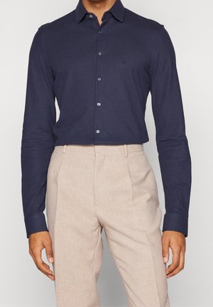 Homme portant une chemise bleu marine cintrée à manches longues boutonnée, rentrée dans un pantalon beige sur mesure et taille haute.