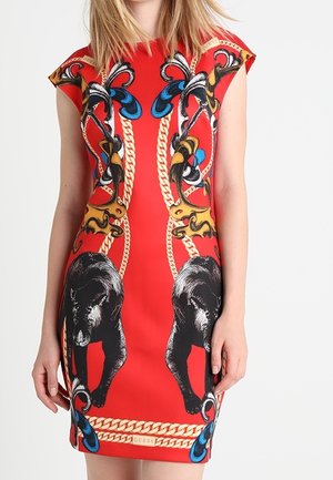 Kobieta w czerwonej dopasowanej sukience z motywem czarnej pantery, złotym łańcuchem i niebieskimi kwiatowymi wzorami, stojąca na jednolitym białym tle.