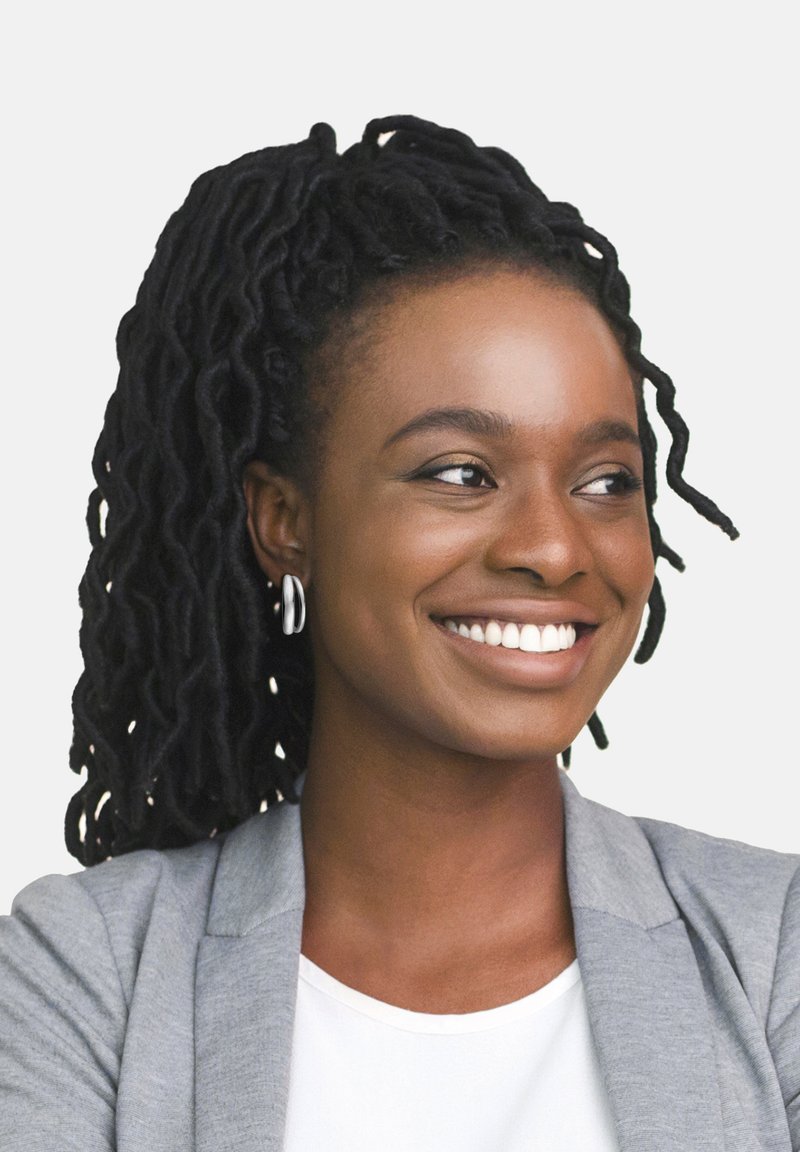 Ritratto di una donna con lunghi dreadlocks neri e texture, che indossa un blazer grigio chiaro sopra una camicia bianca, sorridendo con orecchini a cerchio argento.