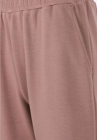 Ljusrosa joggers gjorda av mjukt tyg, med elastisk midja och sidficka, samt en slät, något texturerad yta.