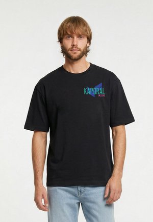 Junger Mann mit hellbraunen Haaren und Bart, der ein schwarzes Kaporal T-Shirt und hellblaue Jeans trägt, vor einem schlichten Hintergrund stehend.
