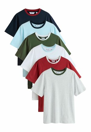 Bărbat tânăr cu păr blond scurt, purtând un tricou gri deschis cu guler întunecat și blugi închiși la culoare, cu mâinile încrucișate, arătând tatuajele de pe antebrațe.