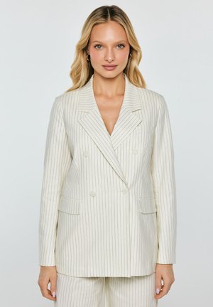 Blondynka w białej, prążkowanej, dwurzędowej marynarce z dopasowanymi spodniami, stojąca na tle jasnego, jednolitego tła.