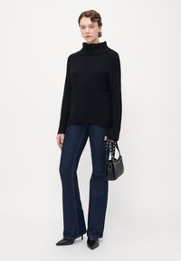 Kvinna iklädd en svart polotröja, mörkblå jeans, svarta spetsiga klackar och håller en svart nitas handväska framför en neutral bakgrund.