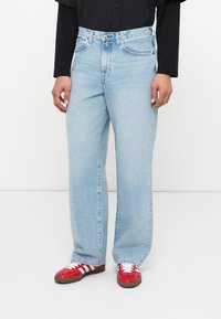 Jeans de ganga azul claro com corte reto, dois bolsos frontais e fecho de botão, combinados com ténis vermelhos com detalhes brancos.