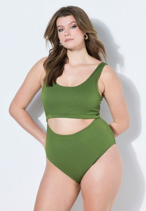 Mujer con el pelo largo y castaño, vestida con un bañador verde de dos piezas con textura acanalada, de pie con la mano en la cadera sobre fondo blanco.