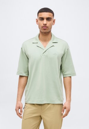 Muž s krátkymi vlasmi, oblečený v svetlozelenej košeli s krátkym rukávom a golierom a béžových nohaviciach, stojaci pred jednoduchým bielym pozadím.