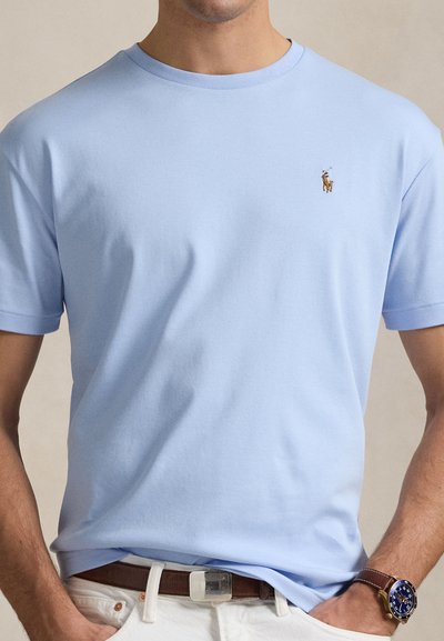 Šviesiai mėlynas medvilninis marškinėlis su apvaliu iškirpte ir trumpomis rankovėmis; ant krūtinės yra mažas siuvinėtas logotipas su arkliu ir raiteliu.