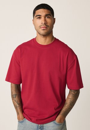 Bărbat cu păr scurt și tatuaje, purtând un tricou roșu supradimensionat și blugi albastru deschis, stând cu mâinile în buzunare.