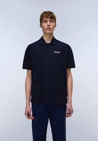 Ung mann med kort krøllete hår iført en mørk marineblå poloskjorte med en liten logo på brystet, stående foran en ensfarget lys bakgrunn.