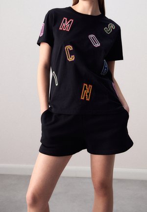 Persona con camiseta de manga corta negra con letras bordadas de colores y pantalones cortos negros a juego, de pie con las manos en los bolsillos contra un fondo liso.
