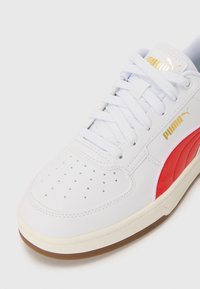 Vit sneaker med en slät läderöverdel, röda accenter och guldlogotyp. Den har perforerade detaljer på tån och en gummisula.