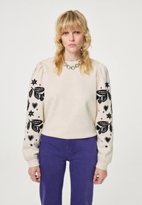 Cremfarbener Sweatshirt mit schwarzen gestickten Blumen- und Sternmotiven auf gepufften Ärmeln. Kombiniert mit lila Hosen, die über einen runden Halsausschnitt verfügen.
