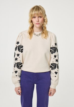 Cream sweatshirt with black embroidered floral and star designs on puffed sleeves. Paired with purple pants, featuring a round neckline.