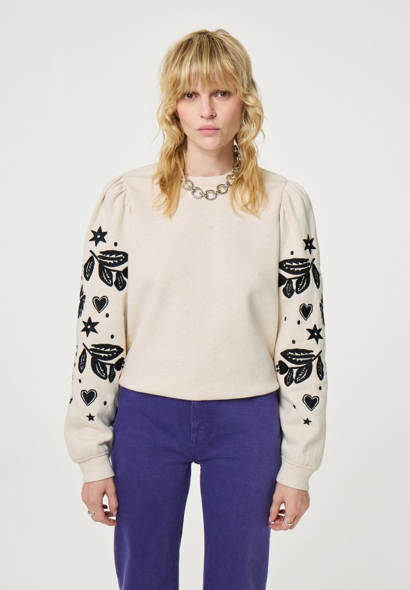 Cremfarbener Sweatshirt mit schwarzen gestickten Blumen- und Sternmotiven auf gepufften Ärmeln. Kombiniert mit lila Hosen, die über einen runden Halsausschnitt verfügen.