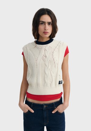 Femme aux longs cheveux bruns portant un pull sans manches en câble tricoté crème sur un t-shirt rouge à manches courtes et un jean bleu foncé, les mains dans les poches.